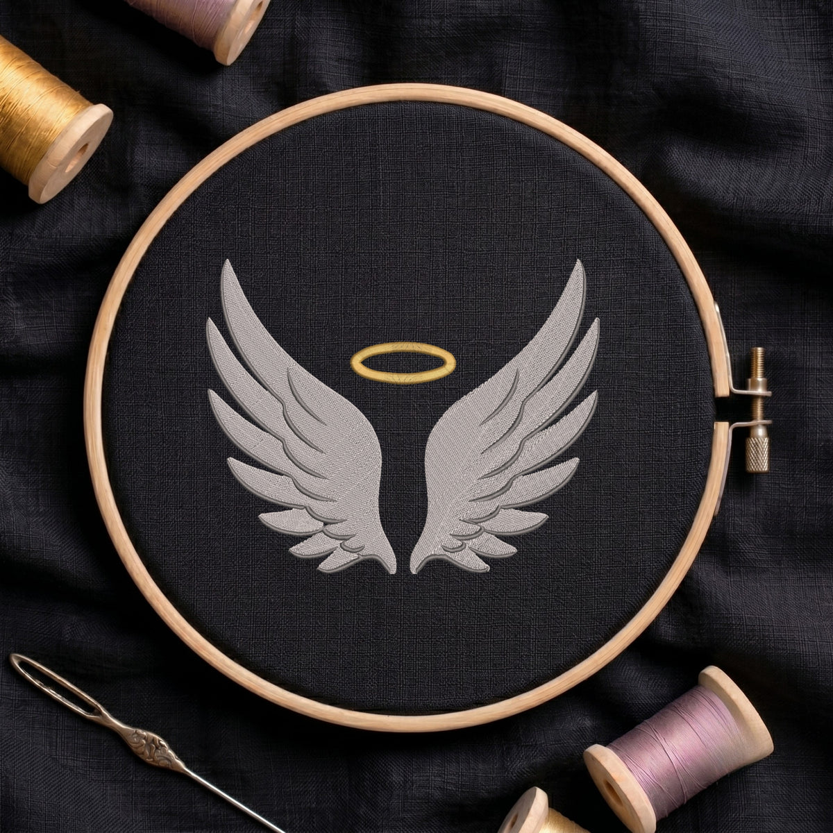 A circular embroidery hoop holding black fabric with a gray angel wings design and a small gold halo, surrounded by spools of thread and a needle on a dark fabric background.