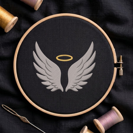 A circular embroidery hoop holding black fabric with a gray angel wings design and a small gold halo, surrounded by spools of thread and a needle on a dark fabric background.