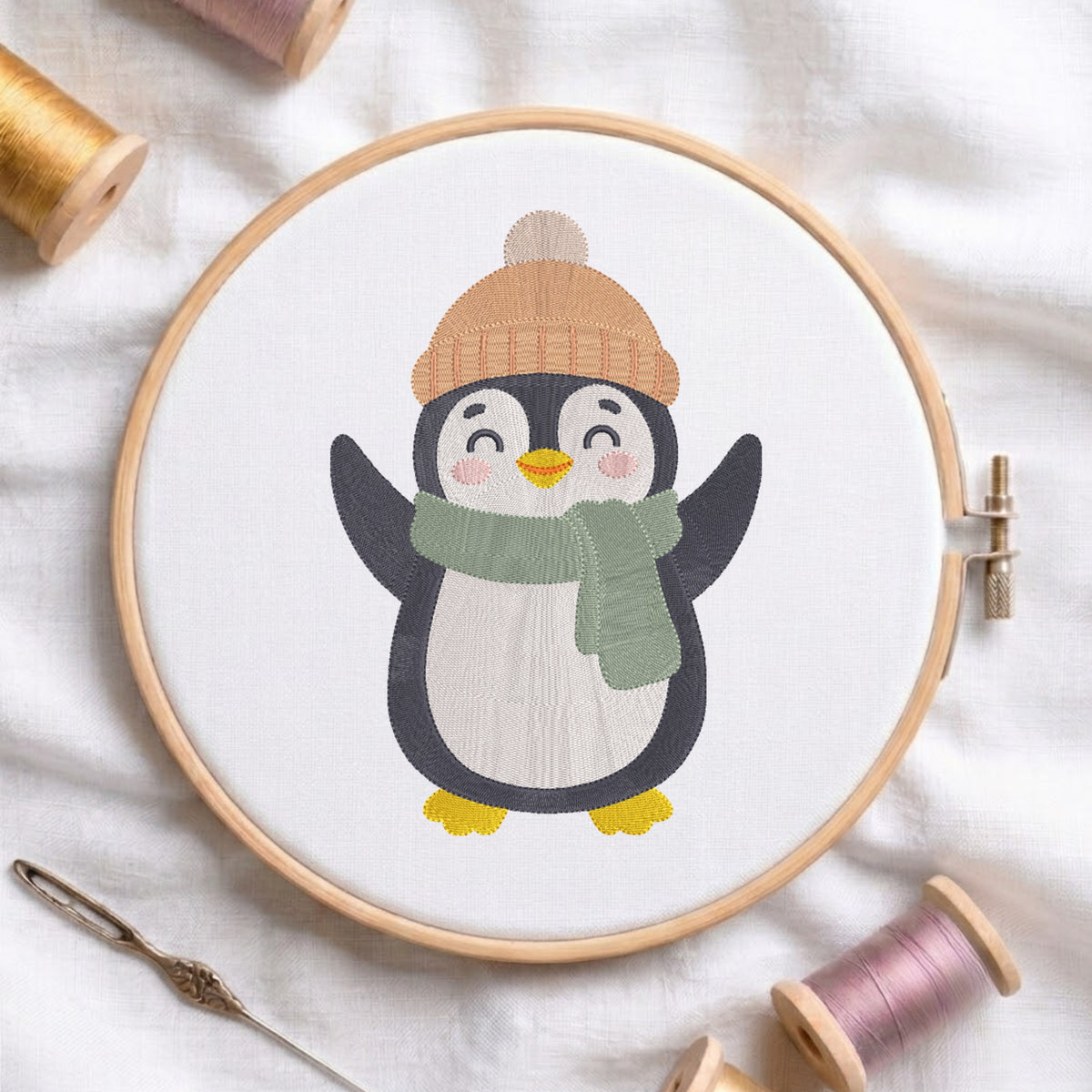 A wooden embroidery hoop holds a white fabric with a stitched penguin design wearing a tan knit hat and green scarf, surrounded by spools of thread and a needle on a white fabric background.