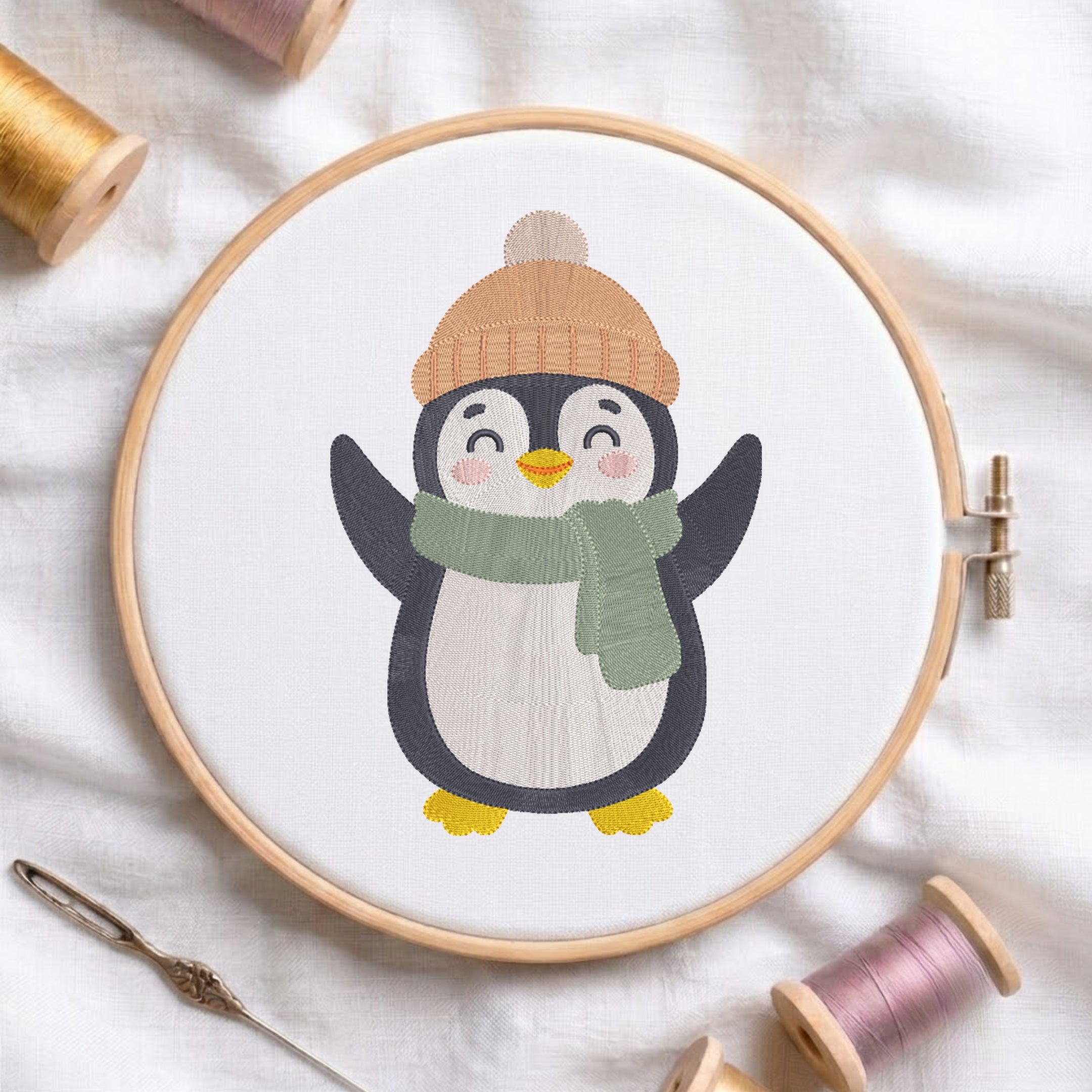 A wooden embroidery hoop holds a white fabric with a stitched penguin design wearing a tan knit hat and green scarf, surrounded by spools of thread and a needle on a white fabric background.