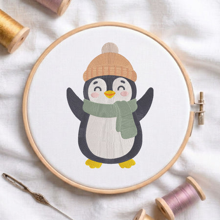 A wooden embroidery hoop holds a white fabric with a stitched penguin design wearing a tan knit hat and green scarf, surrounded by spools of thread and a needle on a white fabric background.