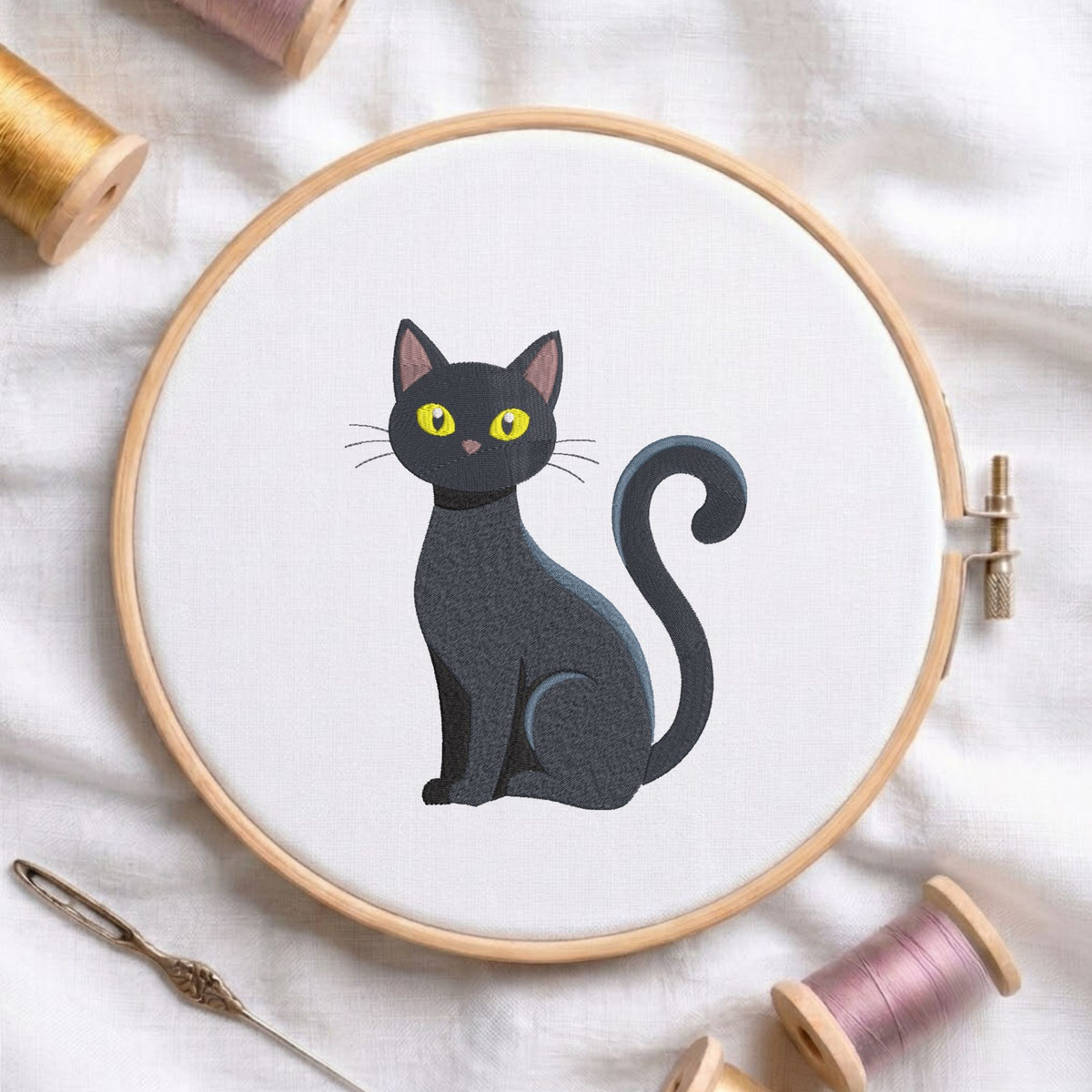 Cute sitting black cat with big yellow eyes and a curved tail machine embroidery design, displayed inside a round wooden hoop on white fabric with thread spools and a needle.