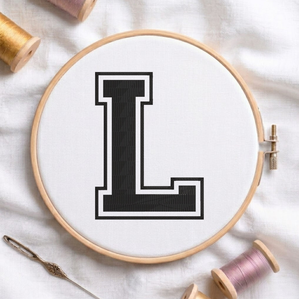 A wooden embroidery hoop holding white fabric with a large black letter L outlined in white, surrounded by spools of thread and a needle on a white fabric background.