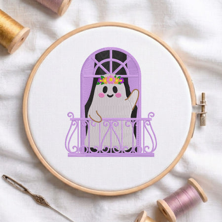 A circular embroidery hoop holding a white fabric panel with a cartoon ghost character waving from behind a purple balcony and window frame. The hoop is photographed on a white fabric background with spools of thread and a needle around it.