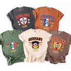Five t-shirts in charcoal, rust, green, cream, and brown colors laid out flat, each printed with a retro crest design featuring a soccer ball, country flag themes, and the year 2026.