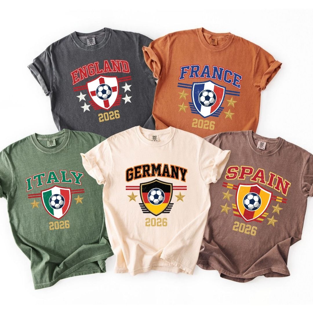 Five t-shirts in charcoal, rust, green, cream, and brown colors laid out flat, each printed with a retro crest design featuring a soccer ball, country flag themes, and the year 2026.