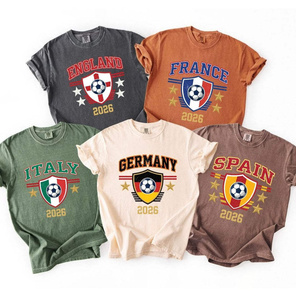Five t-shirts in charcoal, rust, green, cream, and brown colors laid out flat, each printed with a retro crest design featuring a soccer ball, country flag themes, and the year 2026.