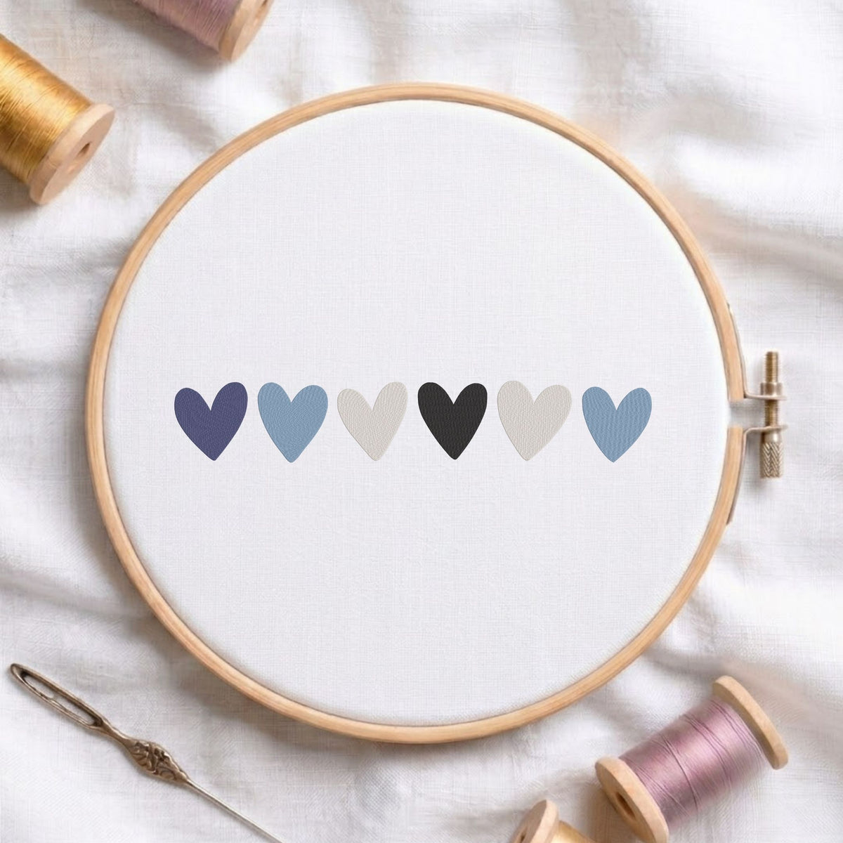 A wooden embroidery hoop holding white fabric with six small hearts in muted blue, gray, black, and beige tones. The hoop is styled on a white fabric background with spools of thread and a needle nearby.
