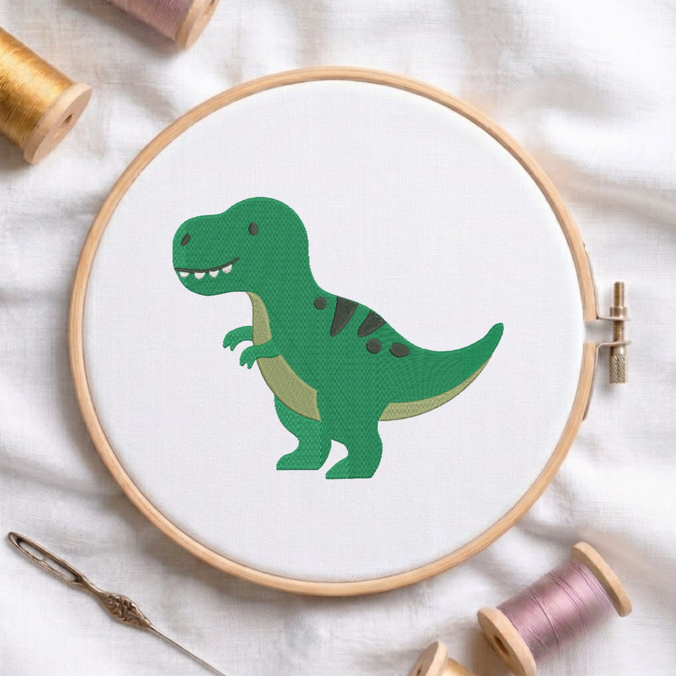 A wooden embroidery hoop holding white fabric with a green cartoon dinosaur design, surrounded by thread spools and a needle on a white fabric background.