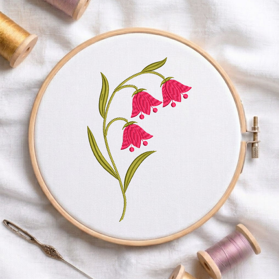 A wooden embroidery hoop holds white fabric with a floral design of pink bell-shaped flowers and green leaves. Surrounding the hoop are spools of thread, a needle, and soft fabric on a light background.