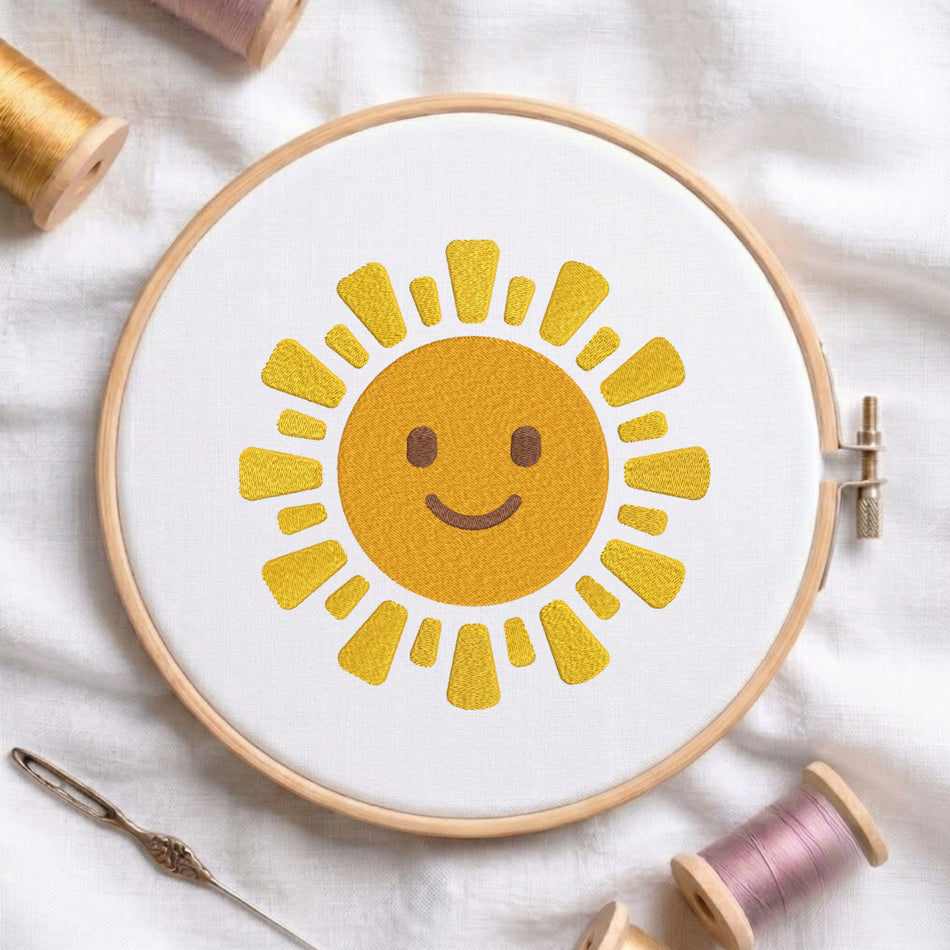 A wooden embroidery hoop holding white fabric with a yellow smiling sun design, surrounded by spools of thread and a needle on a white fabric background.