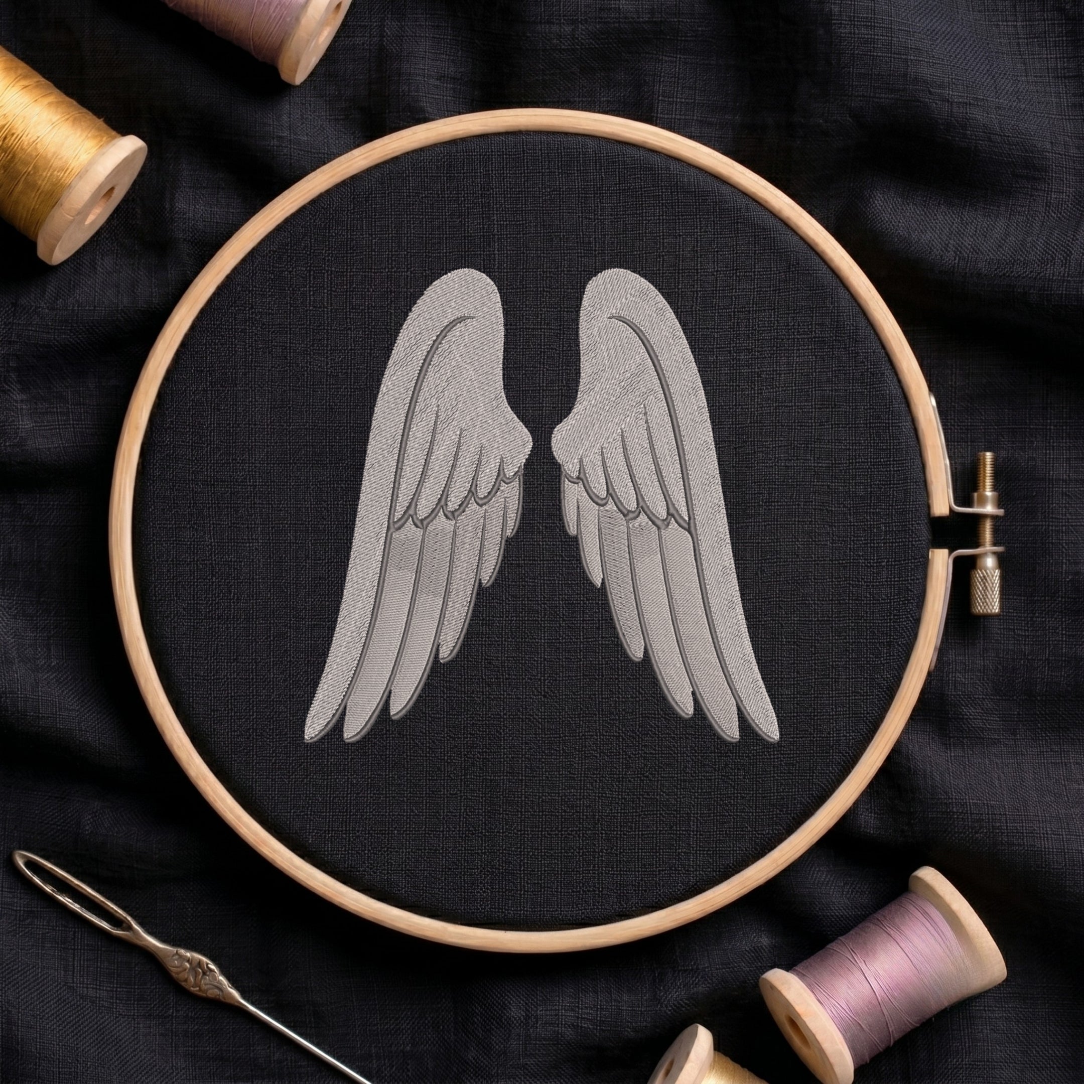 A wooden embroidery hoop holds black fabric with a stitched gray angel wings design in the center. Spools of thread, a needle, and dark fabric are arranged around it in a styled flat-lay.