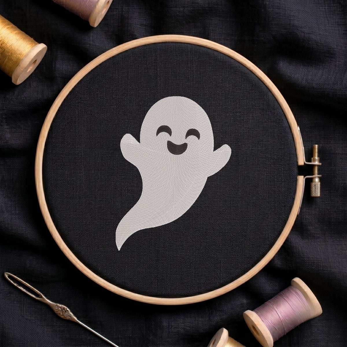 Cute smiling ghost machine embroidery design displayed inside a round wooden hoop on black fabric alongside thread spools and a needle