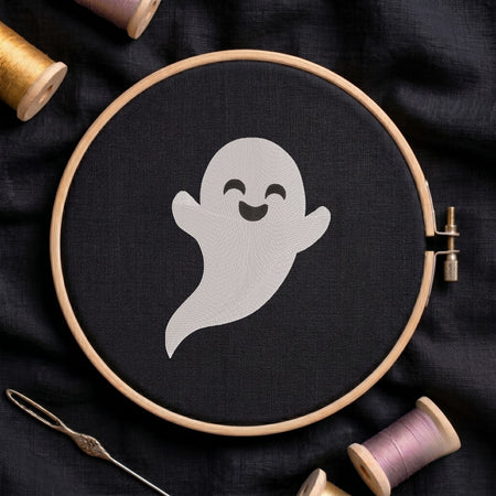 Cute smiling ghost machine embroidery design displayed inside a round wooden hoop on black fabric alongside thread spools and a needle