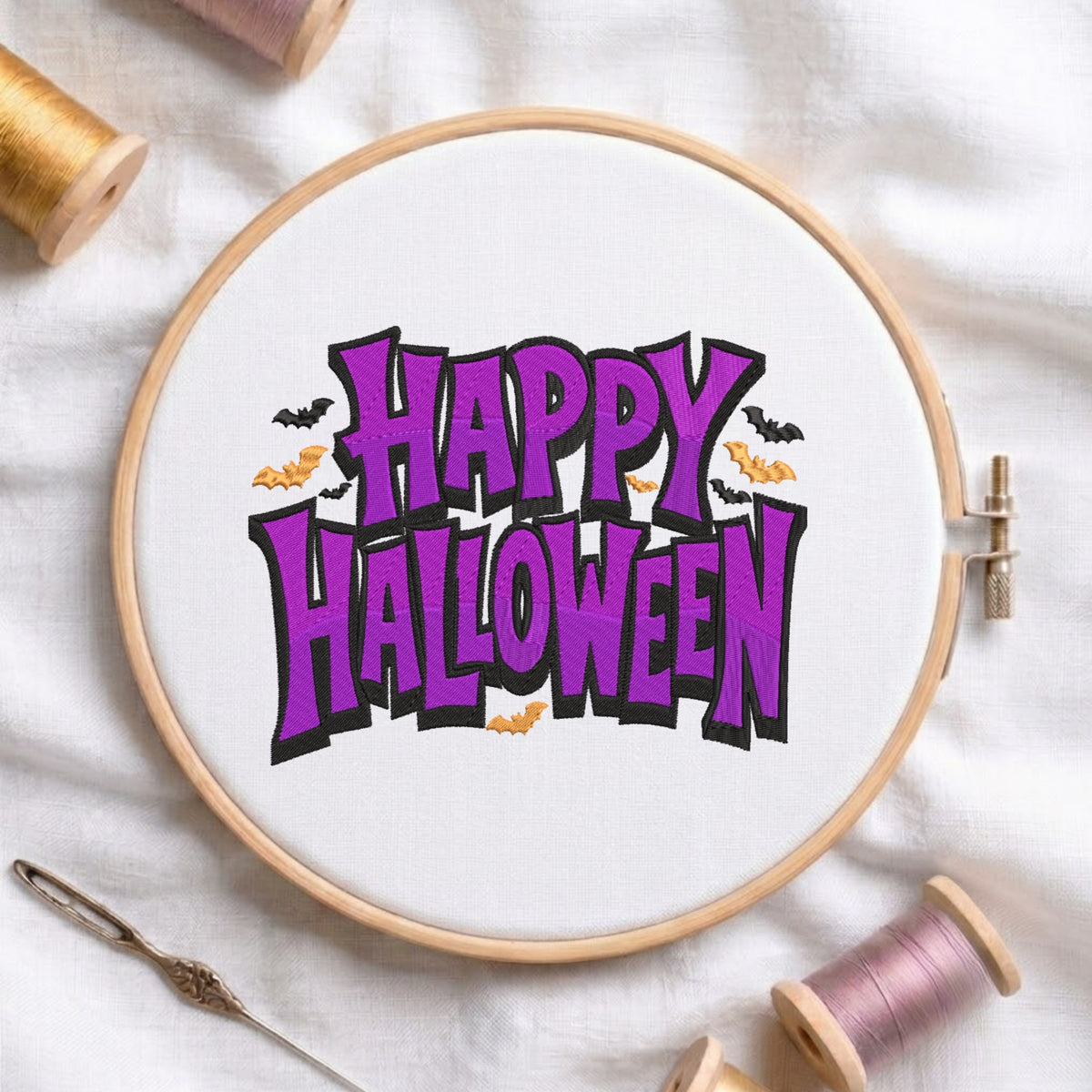 A wooden embroidery hoop holding white fabric with a colorful 'Happy Halloween' design in purple, black, and orange. Spools of thread and a needle are arranged around it on a white fabric background.