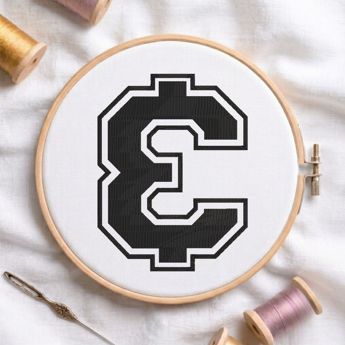 A wooden embroidery hoop holding white fabric with a large black-and-white letter C motif, styled on a white cloth with thread spools and a needle around it.
