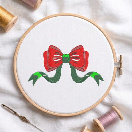 A circular embroidery hoop on white fabric, featuring a red bow with green ribbon tails stitched in the center. Spools of thread and a needle are arranged around it on a white cloth background.