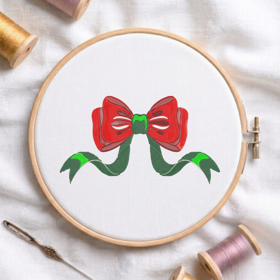 A circular embroidery hoop on white fabric, featuring a red bow with green ribbon tails stitched in the center. Spools of thread and a needle are arranged around it on a white cloth background.