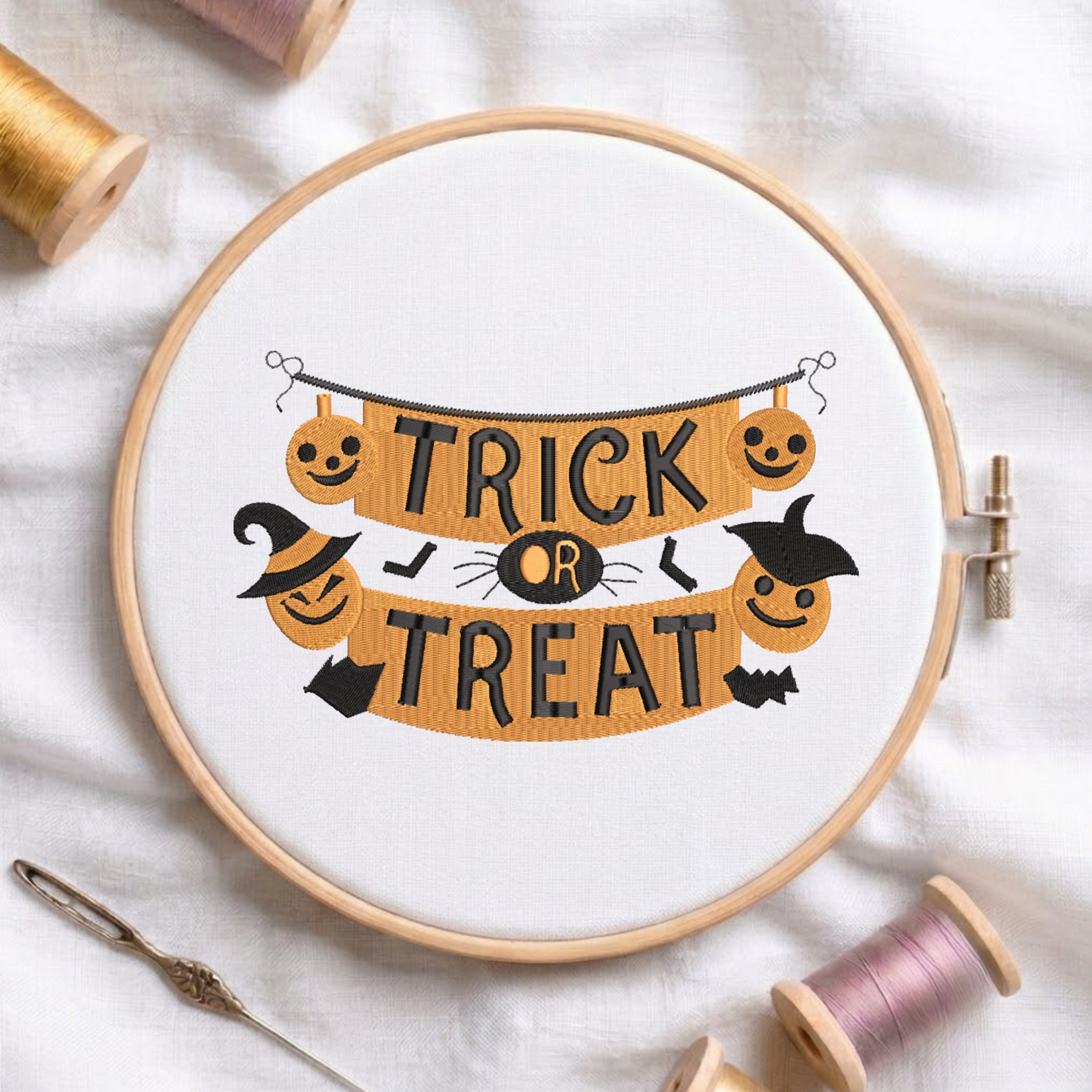 A circular embroidery hoop containing a white fabric with a Halloween-themed cross-stitch design that reads 'TRICK OR TREAT' in black letters over an orange banner with jack-o'-lanterns, a witch hat, bats, and a spider. The hoop is styled on a white fabric background with spools of thread and a needle.