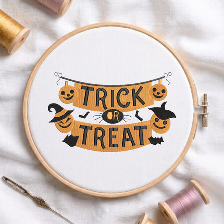 A circular embroidery hoop containing a white fabric with a Halloween-themed cross-stitch design that reads 'TRICK OR TREAT' in black letters over an orange banner with jack-o'-lanterns, a witch hat, bats, and a spider. The hoop is styled on a white fabric background with spools of thread and a needle.