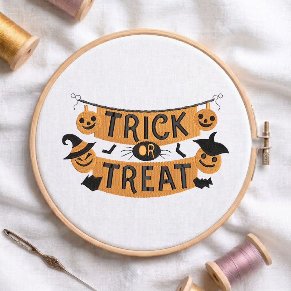 A circular embroidery hoop containing a white fabric with a Halloween-themed cross-stitch design that reads 'TRICK OR TREAT' in black letters over an orange banner with jack-o'-lanterns, a witch hat, bats, and a spider. The hoop is styled on a white fabric background with spools of thread and a needle.
