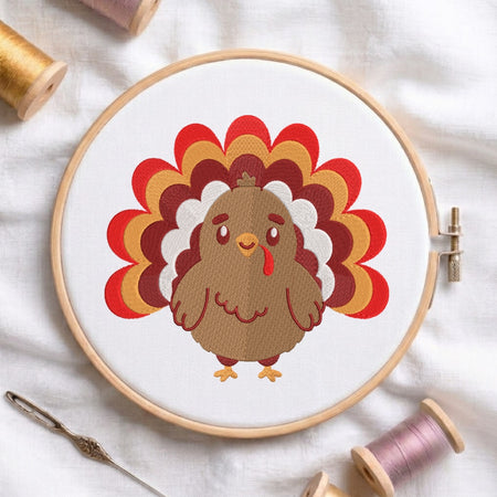 A circular embroidery hoop on white fabric displays a cartoon turkey design with layered red, orange, yellow, and white feathers. Spools of thread and a needle are arranged around the hoop as styled props.