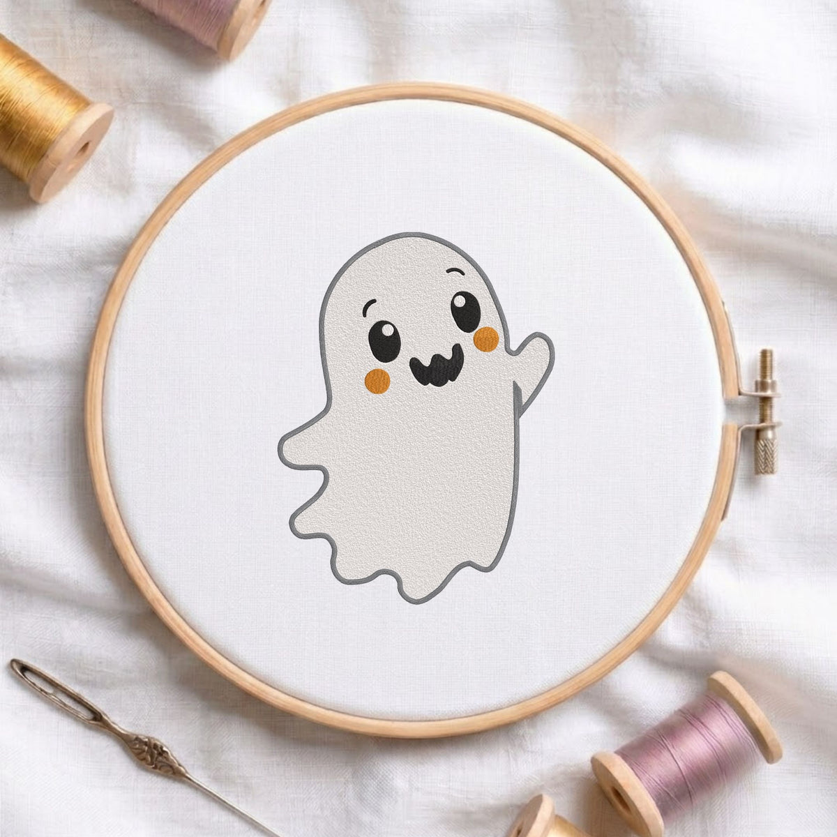 Cute waving ghost machine embroidery design displayed in a wooden embroidery hoop on white fabric with thread spools and a needle