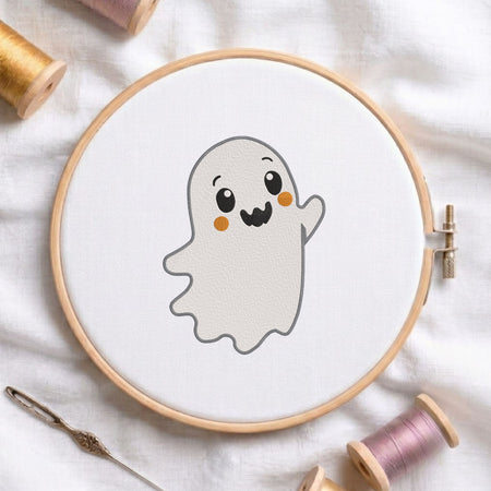 Cute waving ghost machine embroidery design displayed in a wooden embroidery hoop on white fabric with thread spools and a needle