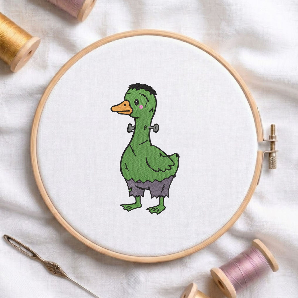 Cute green duck dressed in a Frankenstein monster costume with bolts on its neck for Halloween machine embroidery design shown in a wooden embroidery hoop on white fabric with thread spools and a needle
