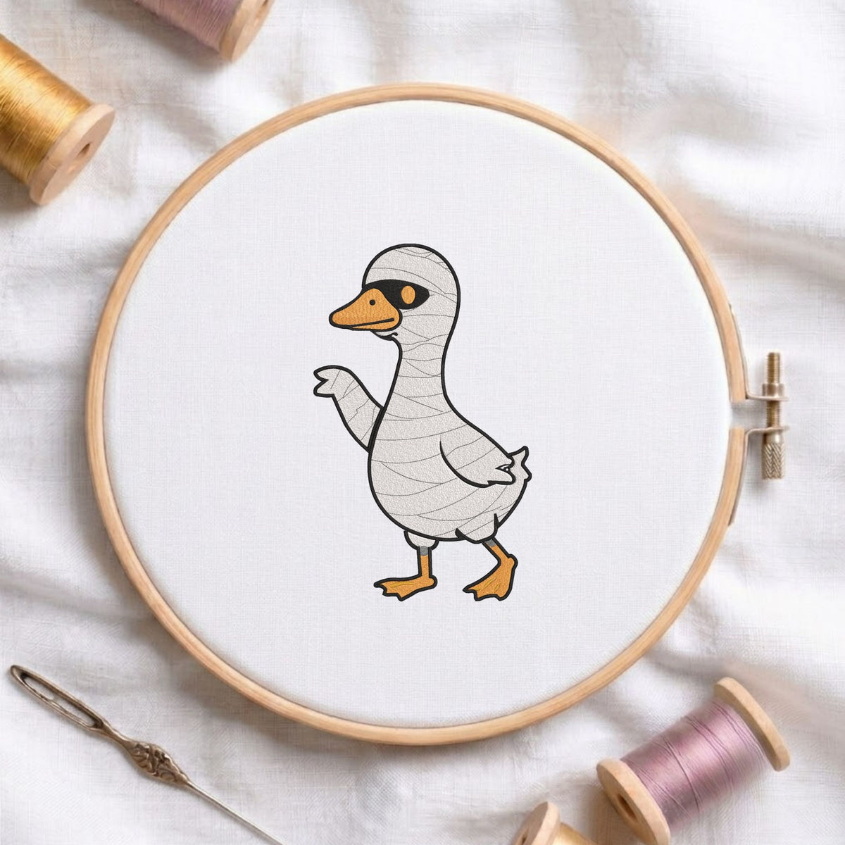Cute duck dressed in a mummy costume for Halloween machine embroidery design shown in a wooden embroidery hoop on white fabric with thread spools and a needle   