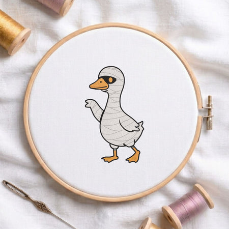 Cute duck dressed in a mummy costume for Halloween machine embroidery design shown in a wooden embroidery hoop on white fabric with thread spools and a needle   