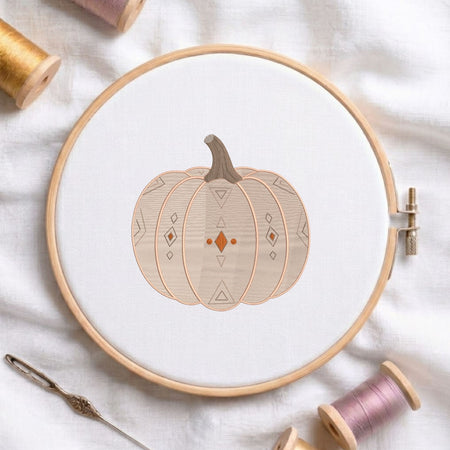 Geometric patterned pumpkin machine embroidery design displayed in a wooden hoop on white fabric with thread spools and a needle.