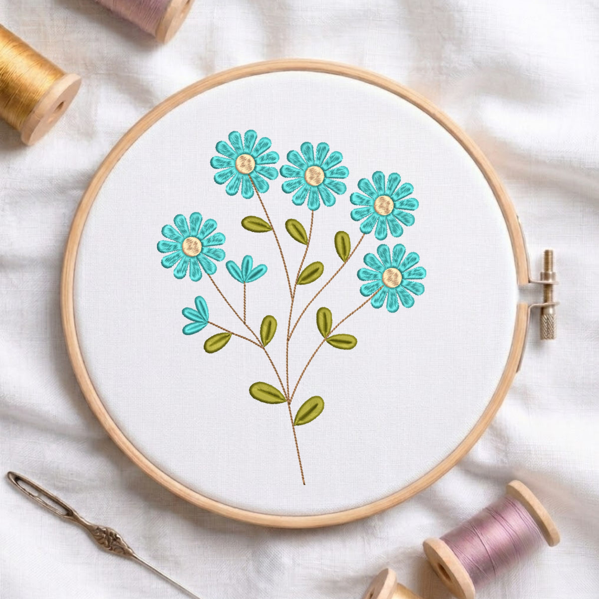 A wooden embroidery hoop holding white fabric with a floral stitched design in turquoise, green, and yellow, surrounded by spools of thread and a needle on a white fabric background.