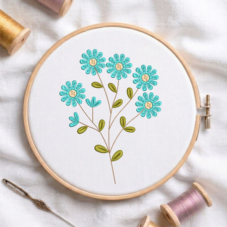 A wooden embroidery hoop holding white fabric with a floral stitched design in turquoise, green, and yellow, surrounded by spools of thread and a needle on a white fabric background.