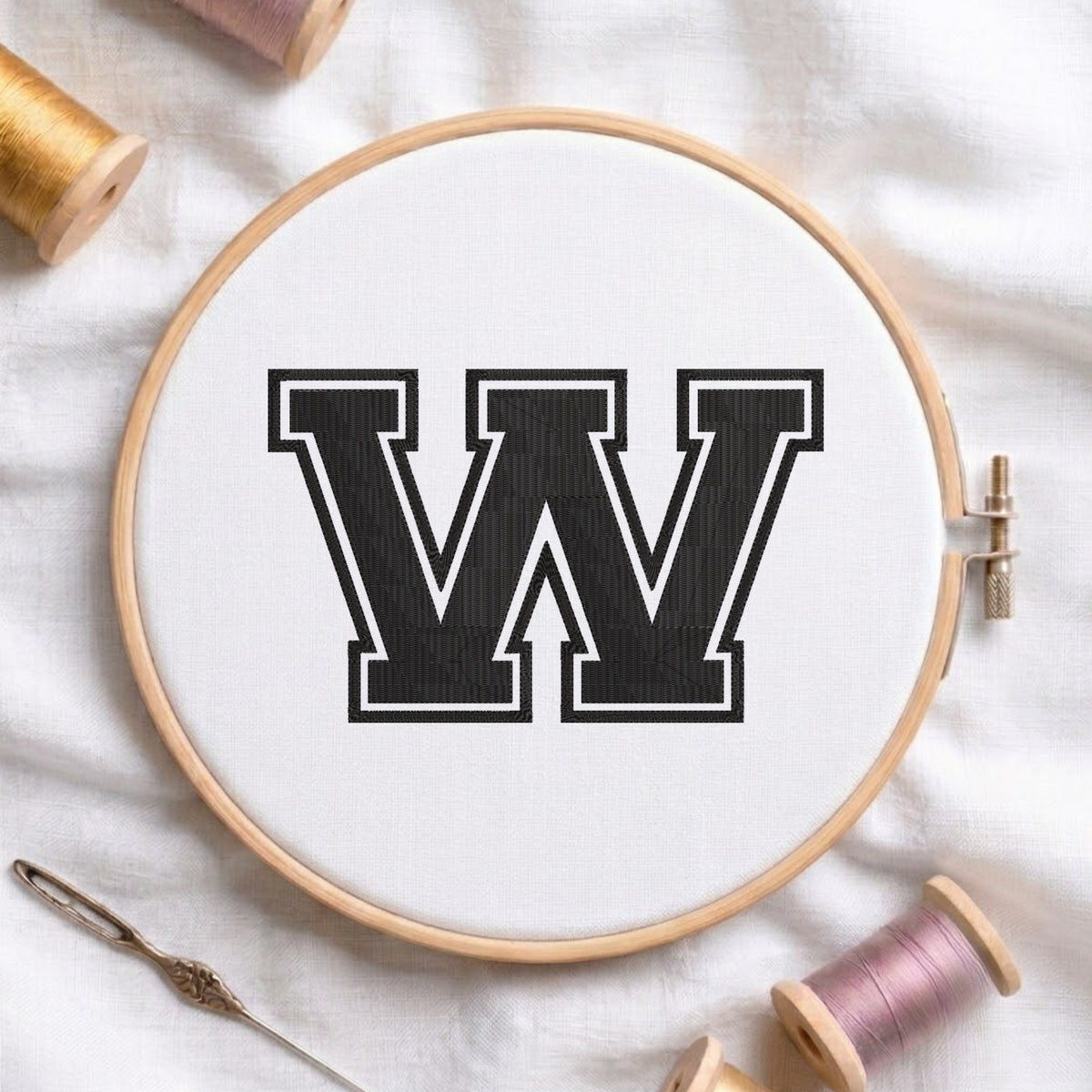 A wooden embroidery hoop containing white fabric with a large black letter W outlined in white, styled on a white fabric background with spools of thread and a needle.