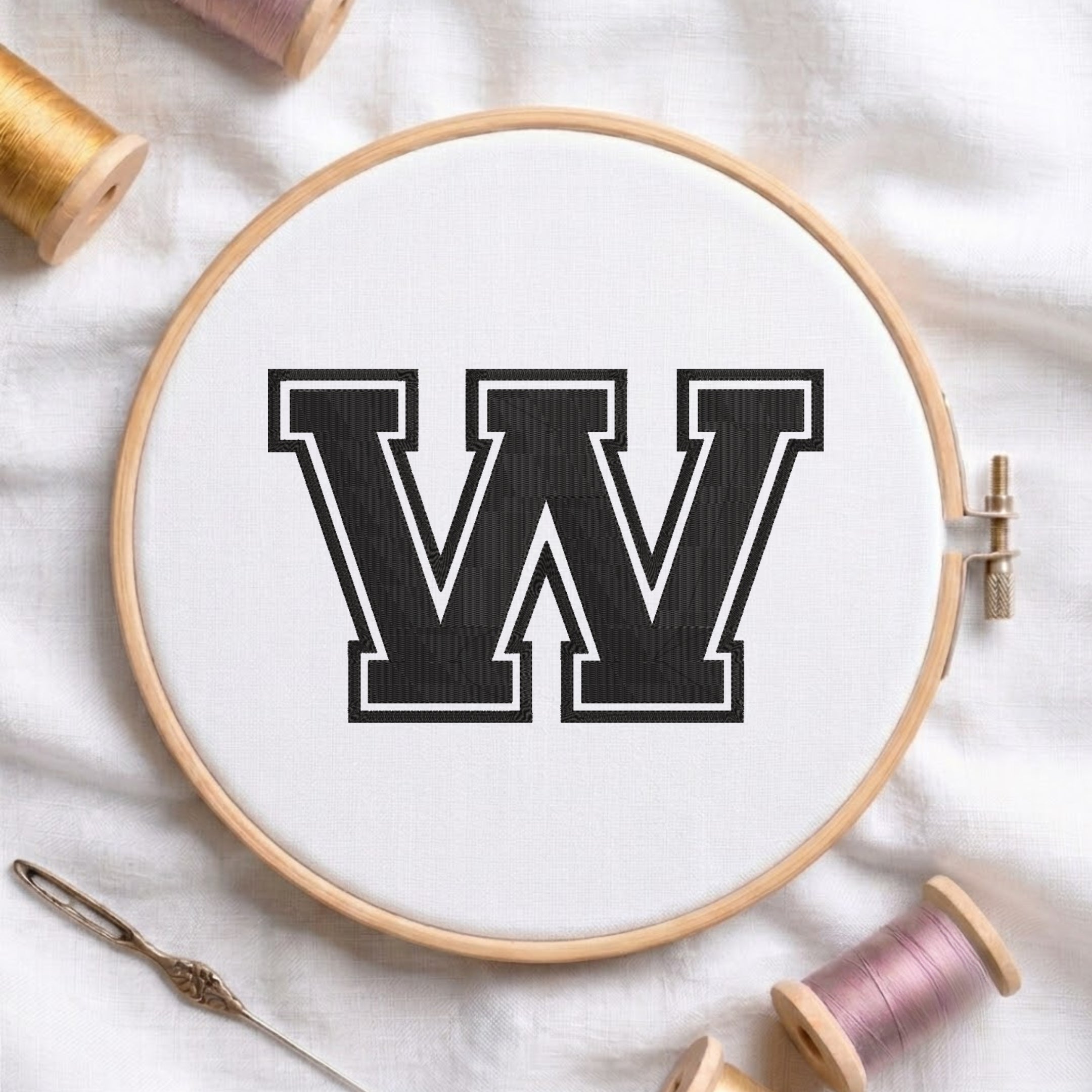 A wooden embroidery hoop containing white fabric with a large black letter W outlined in white, styled on a white fabric background with spools of thread and a needle.