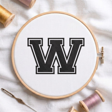 A wooden embroidery hoop containing white fabric with a large black letter W outlined in white, styled on a white fabric background with spools of thread and a needle.