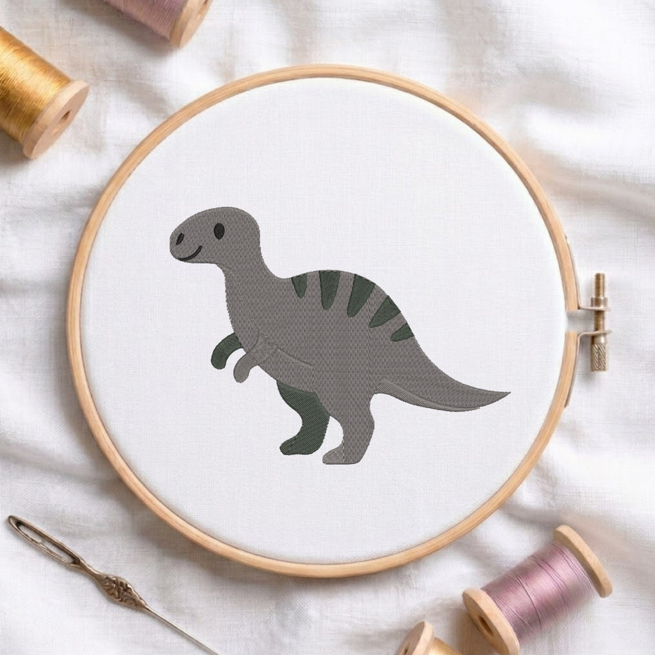 A wooden embroidery hoop holds white fabric with a gray dinosaur design stitched in the center. The hoop is styled on a white fabric background with spools of thread and a needle nearby.