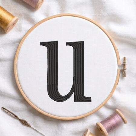 A wooden embroidery hoop holding white fabric with a large black letter 'U' stitched in the center, surrounded by spools of thread and a needle on a white fabric background.