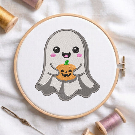 A circular embroidery hoop holding white fabric with a cute ghost design holding a small orange pumpkin. The hoop is styled on a white fabric background with spools of thread and a needle nearby.