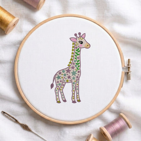 A circular embroidery hoop holding white fabric with a colorful giraffe design, surrounded by spools of thread and a needle on a white fabric background.