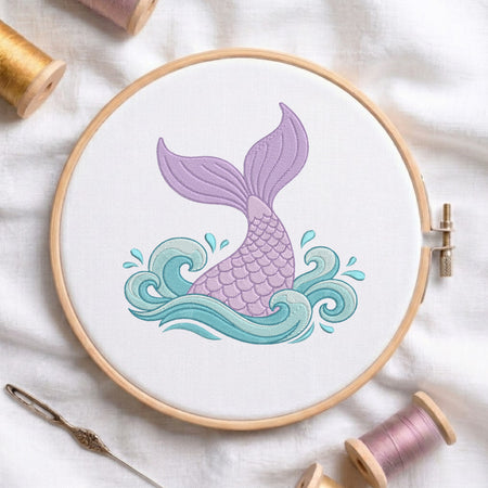 Purple and blue mermaid tail machine embroidery design stitched inside a wooden embroidery hoop on white fabric with spools of thread.