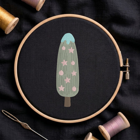 Christmas tree and star ornament machine embroidery design with a green textured body, blue top, and pink star motifs displayed inside a round wooden hoop on black linen fabric, accompanied by wooden thread spools and a needle.