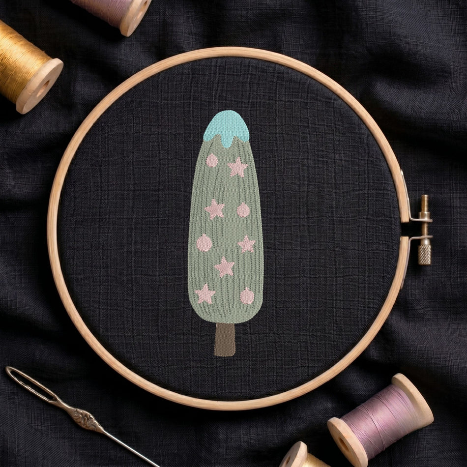 Christmas tree and star ornament machine embroidery design with a green textured body, blue top, and pink star motifs displayed inside a round wooden hoop on black linen fabric, accompanied by wooden thread spools and a needle.