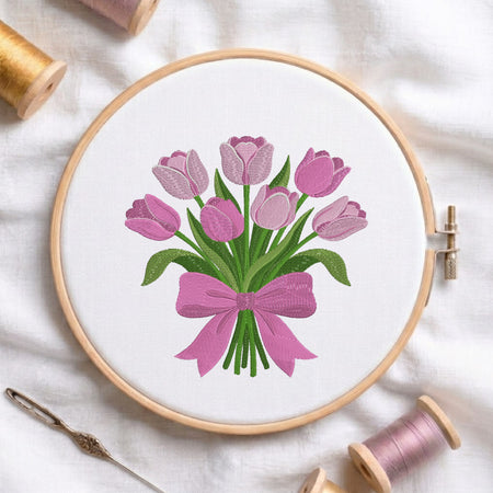 A circular embroidery hoop holds a white fabric with a stitched bouquet of pink tulips and green stems tied with a pink bow. The hoop is photographed on a white fabric surface with spools of thread and a needle around it.
