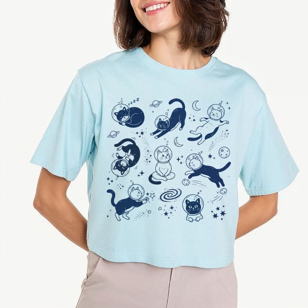 A woman wearing a light blue cropped t-shirt with a dark blue graphic print of cats in space poses against a clean off-white background.