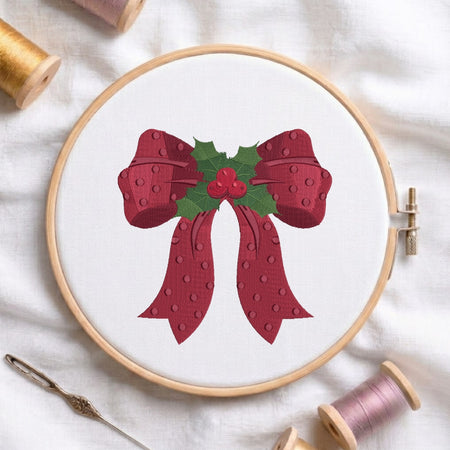 A circular embroidery hoop on white fabric, featuring a festive red bow with green holly leaves and red berries. Spools of thread and a needle are arranged around the hoop in a styled flat-lay composition.