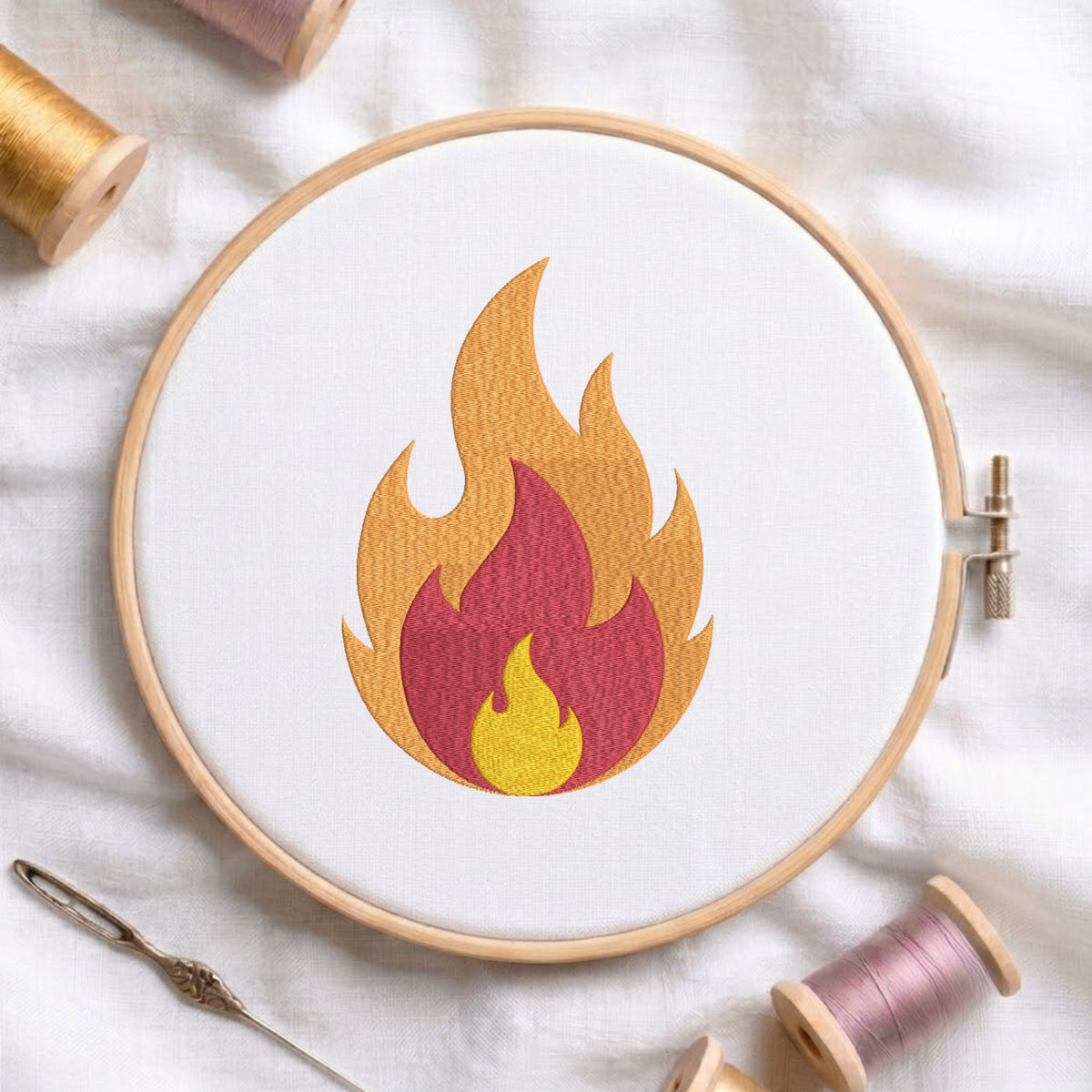 A circular embroidery hoop holds white fabric with a stylized flame motif in yellow, orange, red, and pink. Spools of thread, a needle, and soft fabric are arranged around it in a styled flat-lay.