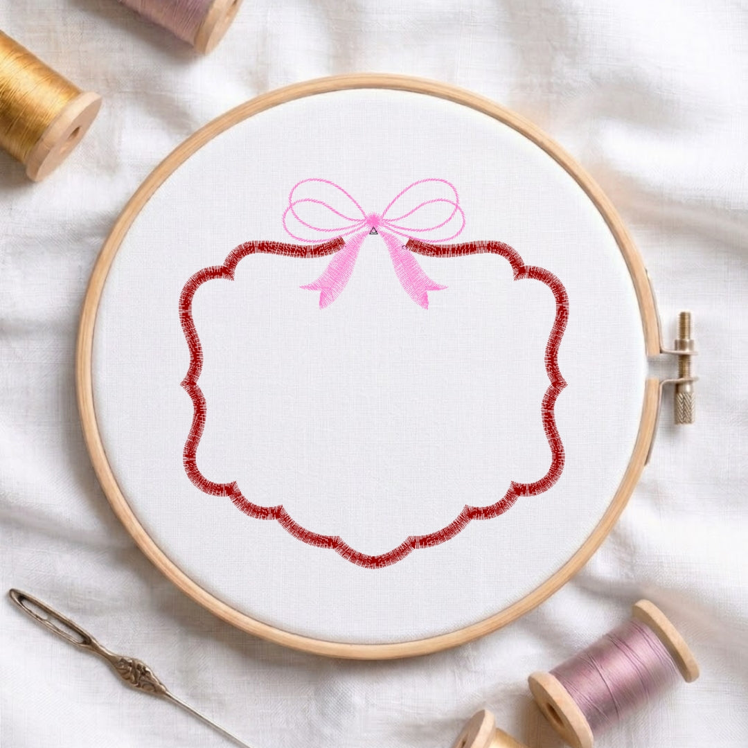 A wooden embroidery hoop holding white fabric with a decorative stitched design: a pink bow above a scalloped red border. Spools of thread, a needle, and a white fabric surface are visible around it.