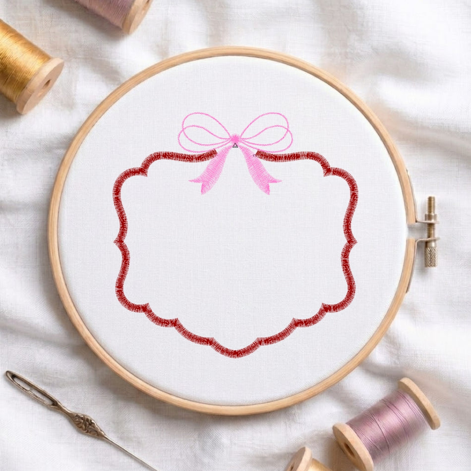 A wooden embroidery hoop holding white fabric with a decorative stitched design: a pink bow above a scalloped red border. Spools of thread, a needle, and a white fabric surface are visible around it.